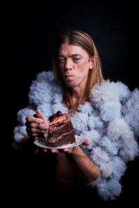 Photo of artist Tom Marshman dressed to party and eating a huge slice of chocolate cake