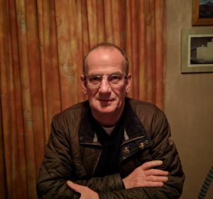 A man is looking straight at the camera. He is wearing glasses and has is arms crossed. He is sitting in front of a curtained background.